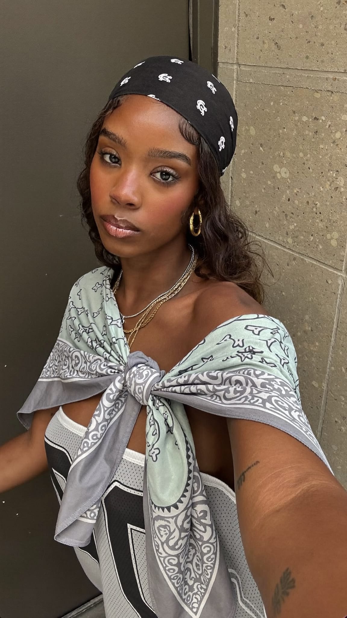 Black woman with waves wearing a large cotton bandana draped and tied in do-rag style, 90s hip-hop aesthetic, oversized jersey and gold jewelry, urban backdrop