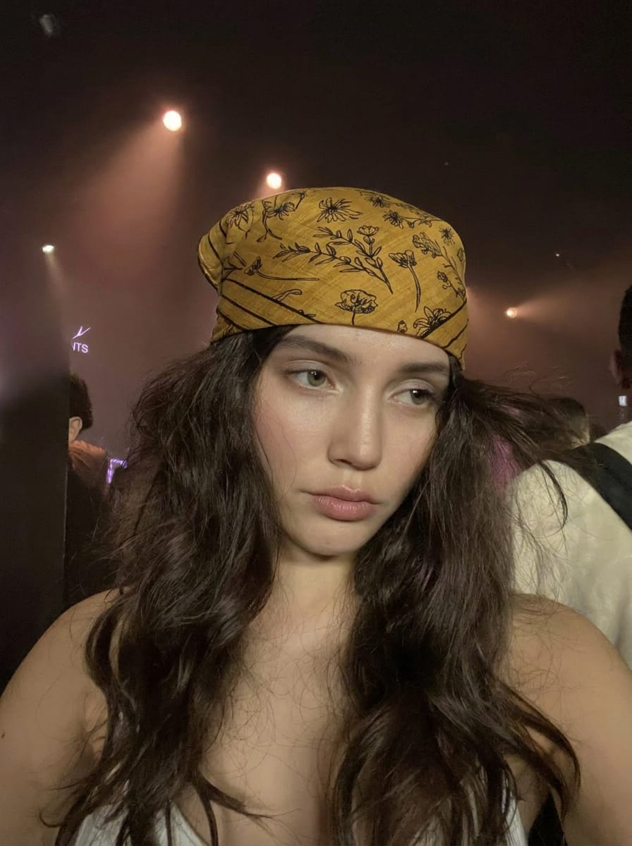 White woman with cool fair skin and messy dark textured hair wearing a printed square bandana tied loosely across the head with an intentionally undone look, moody concert backstage setting with low amber stage lighting and shadows