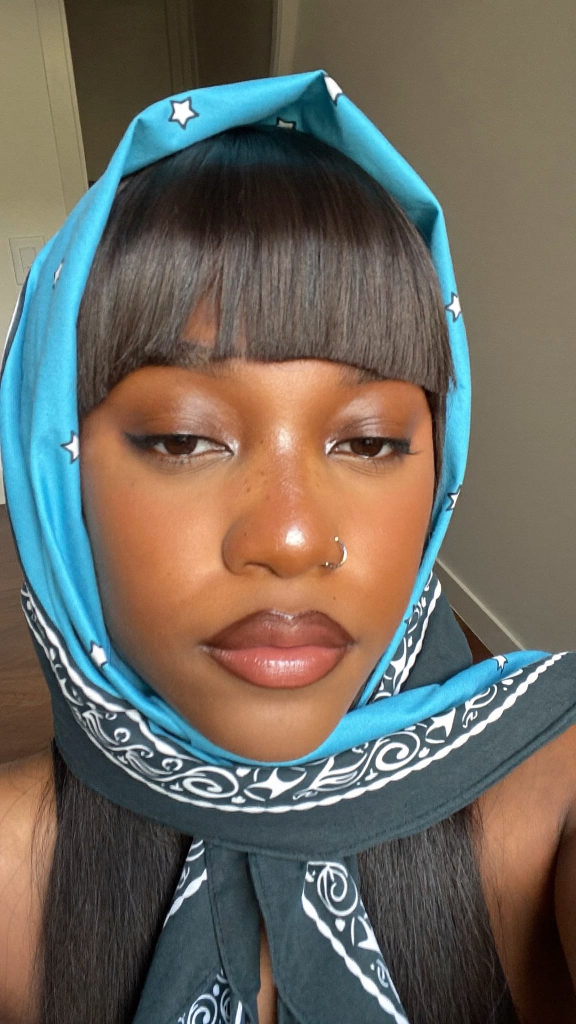 Black woman with blunt even bangs wearing a large cotton bandana wrapped fully around her head with the bangs exposed at the front