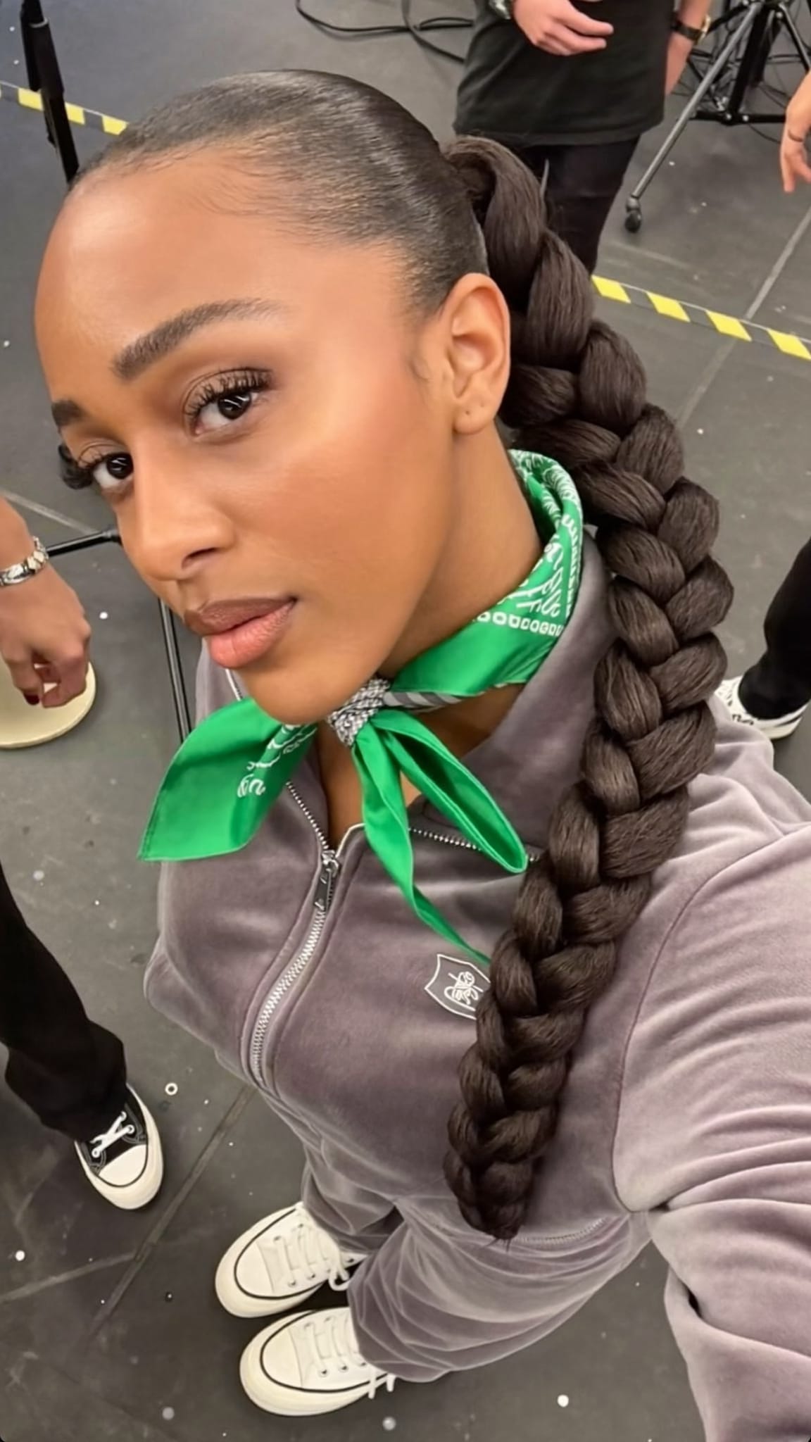 Black woman in her 20s with a sleek high ponytail and silk bandana wrapped around the base, wearing platform sneakers and a velour tracksuit in a music video set