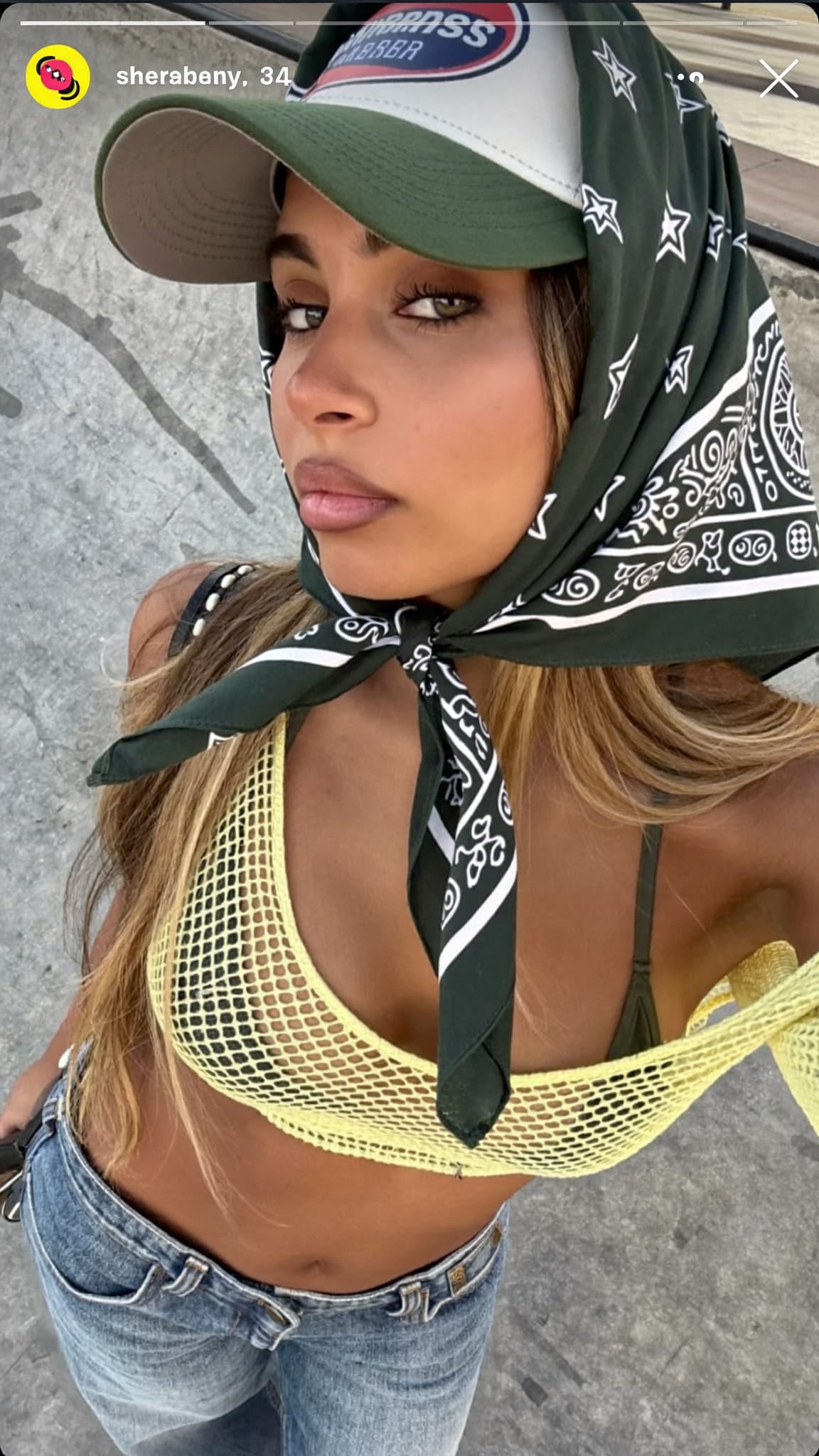 Mixed-race woman in her 20s with a trucker-style cap and a cotton bandana tied at the nape beneath it, wearing a mesh top and low-rise jeans at a skate park