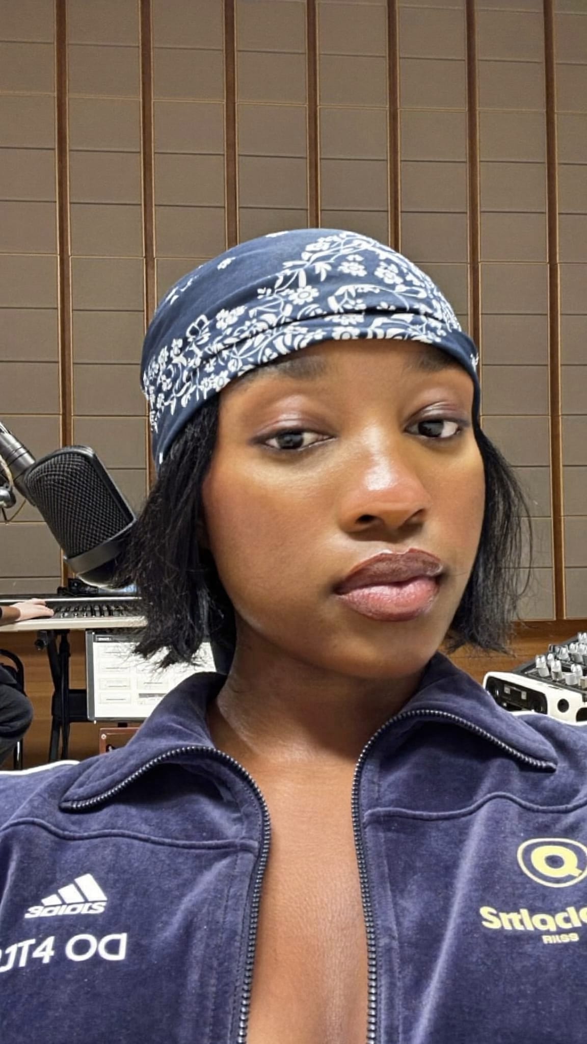 Black woman in her 20s with a short low cut and a navy blue floral bandana worn as a wide headband at the forehead, wearing a velour tracksuit at a recording studio