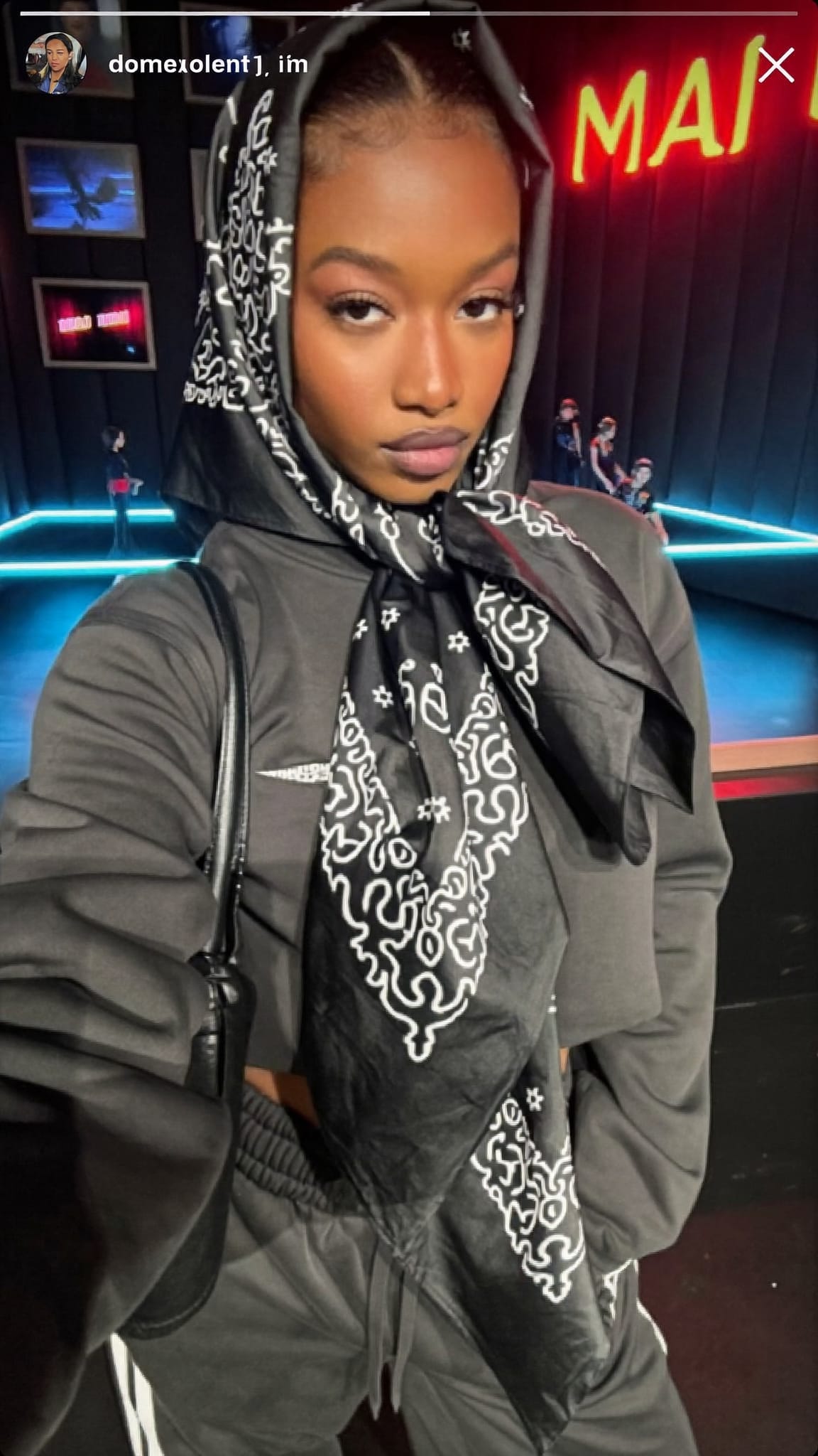 Black woman in her 20s with a bold oversized satin bandana wrapped and tied dramatically at the front, wearing an oversized tracksuit in a neon-lit music video set