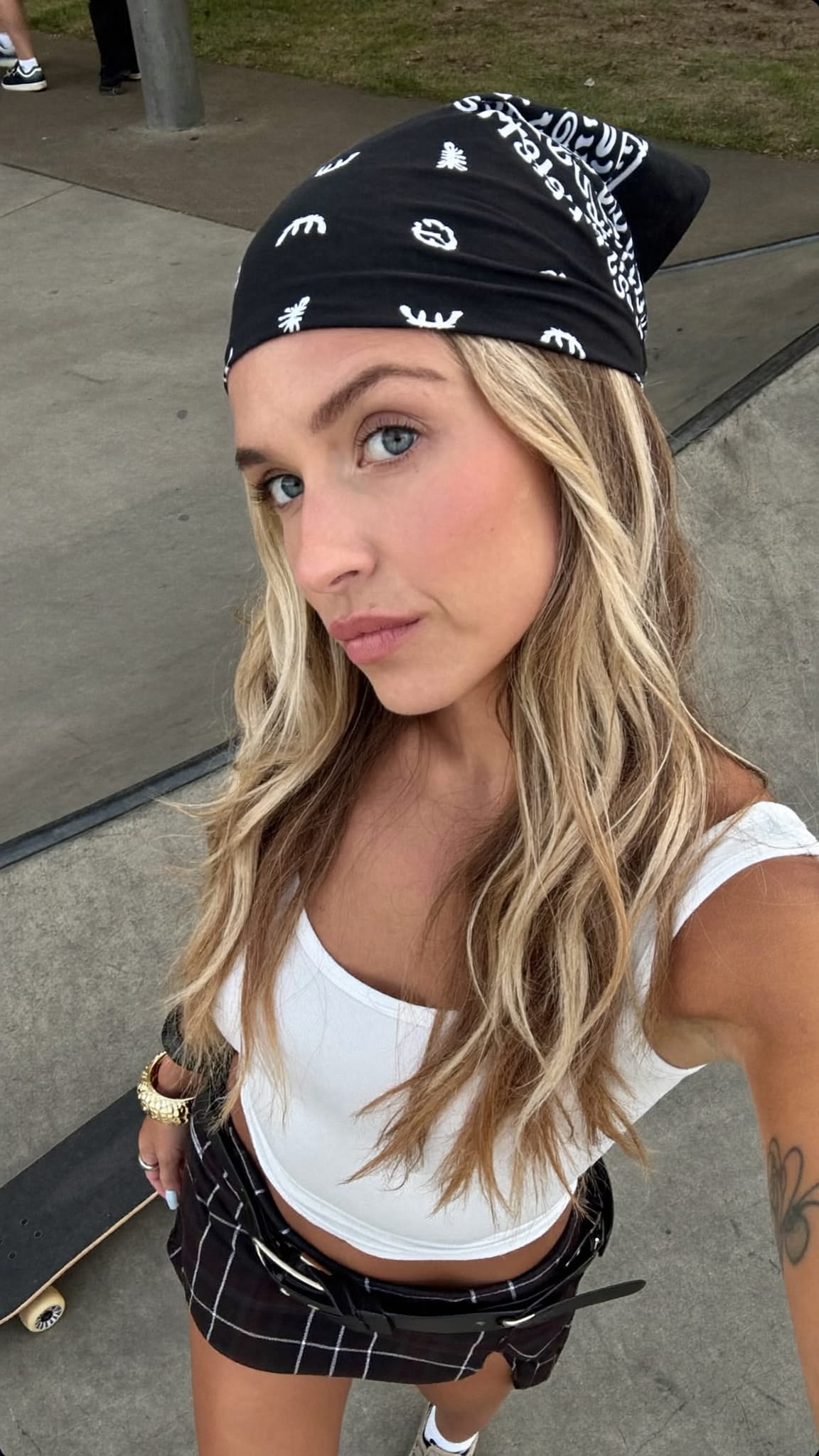 White woman with dirty blonde highlights wearing a cotton bandana as a wide headband with loose hair and a plaid mini skirt and cargo pants at a skate park