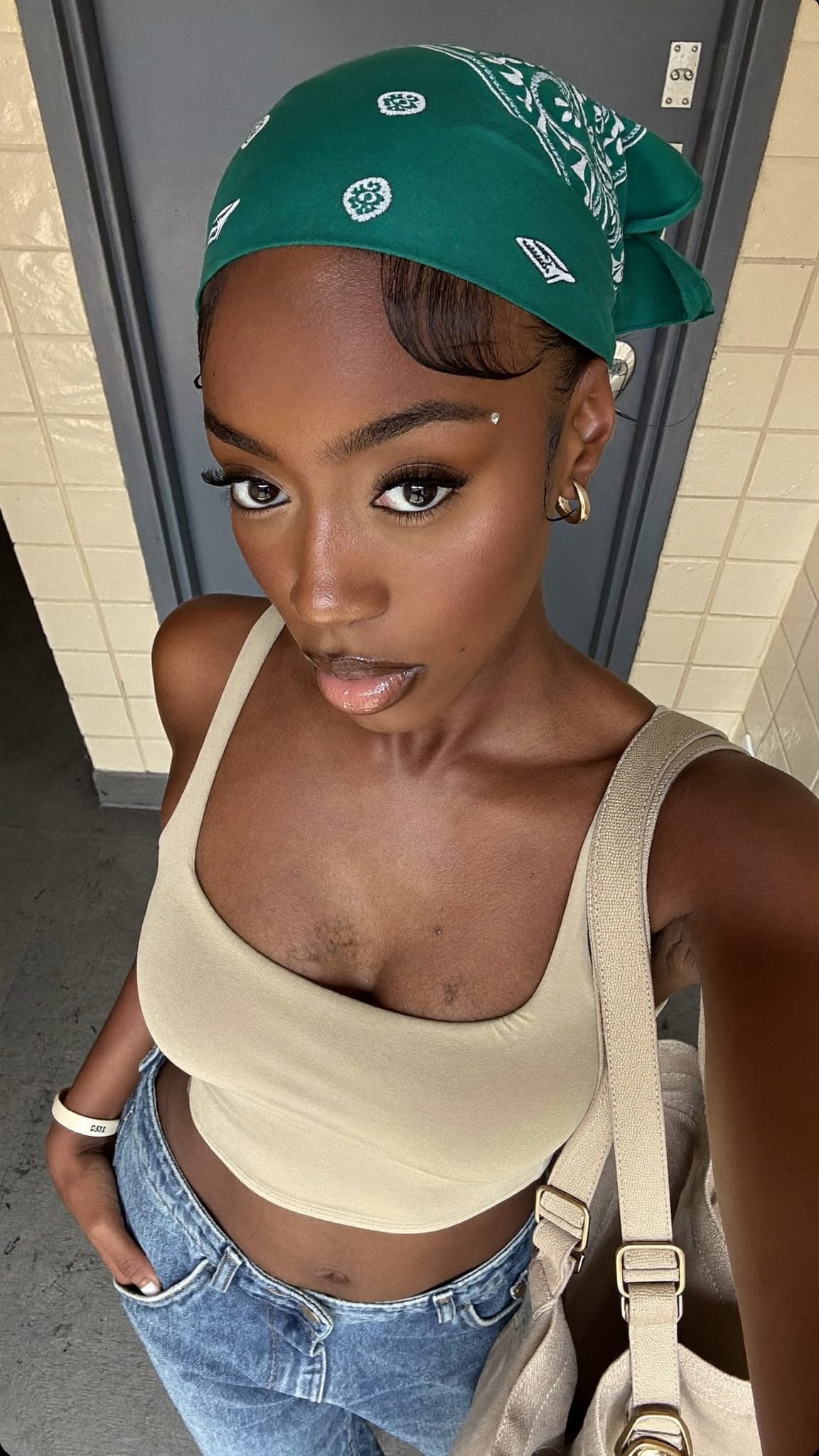 Black woman in her 20s with baby hair edges laid and a cotton bandana headband above the hairline, wearing a crop top and low-rise jeans at a school hallway