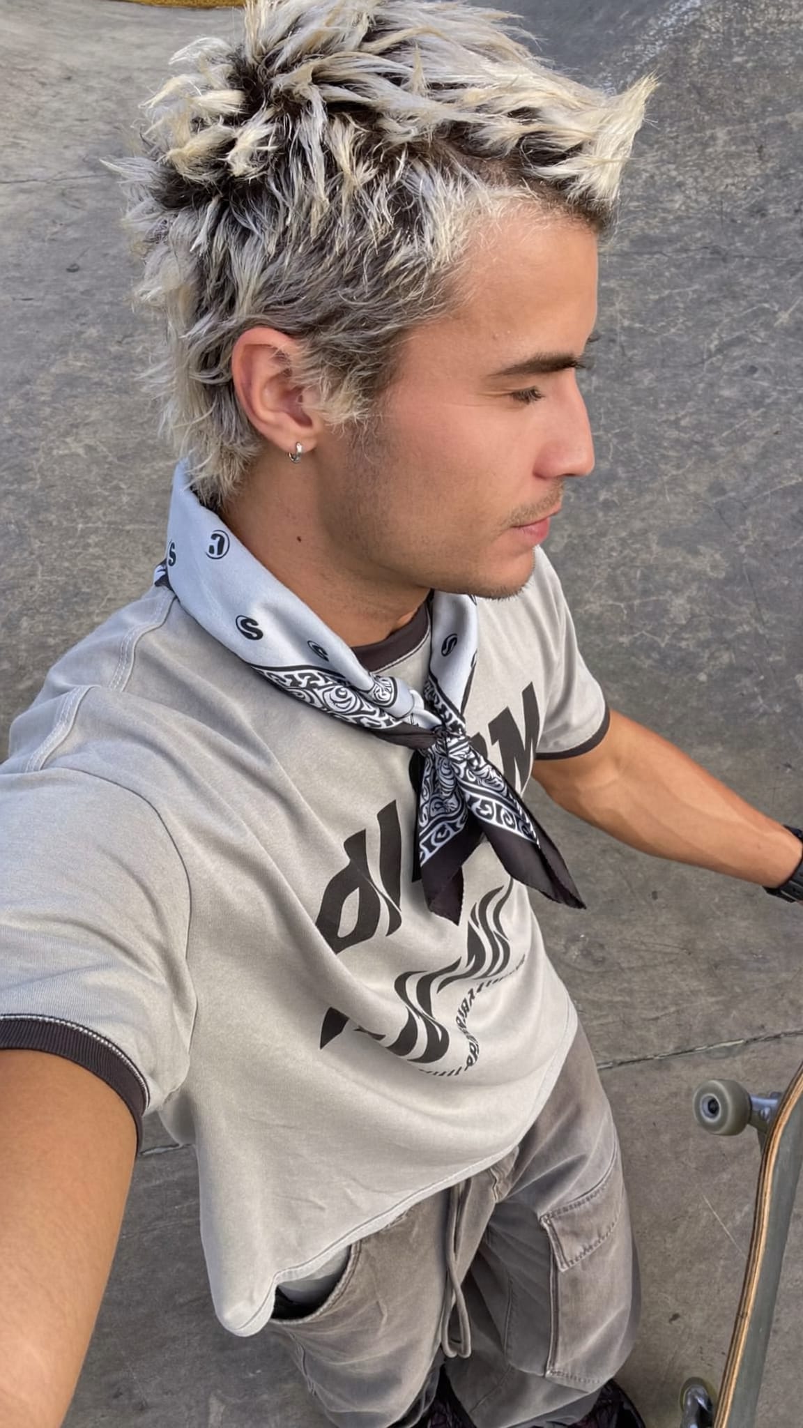 White man in his 20s with frosted-tip spiky hair and a cotton bandana tied loosely at the back of the neck, wearing a skater tee and baggy cargo pants at a skate park