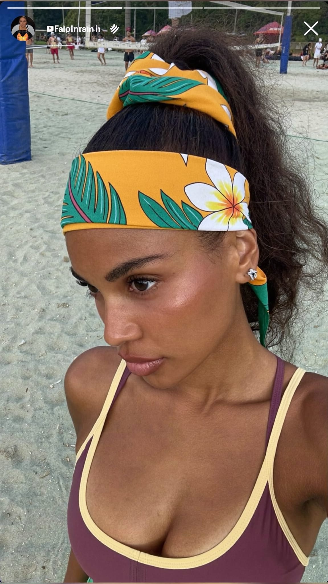 South Asian woman in her 20s with a high athletic ponytail and a tropical island-print bandana tied as a sun-blocking headband, wearing a sports bikini at a beach volleyball court