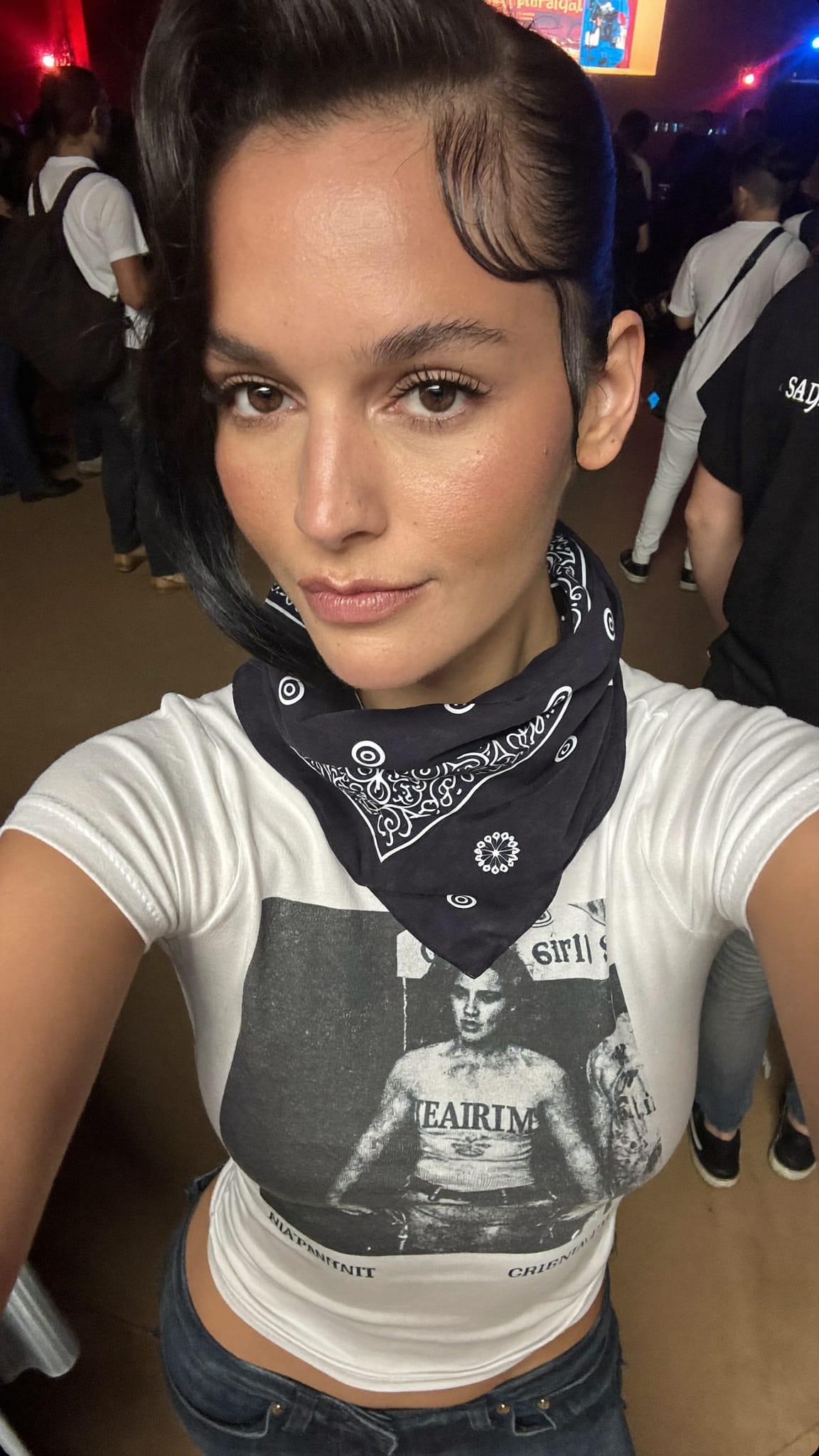 White woman in her early 20s with dark side-swept scene hair and a dark grunge bandana wrapped around the forehead, wearing a band tee and skinny jeans at a concert venue
