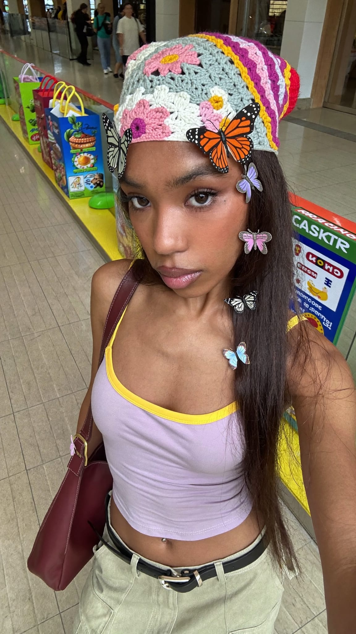 Mixed-race woman in her 20s with butterfly clips scattered in hair and a colorful crochet daisy bandana headband, wearing a pastel crop top and cargo pants at a mall food court