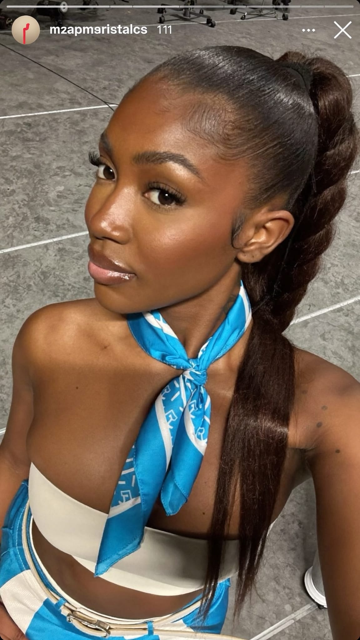 Black woman in her 20s with a high sleek ponytail and a satin bandana tied at the base with long tails, wearing a matching coordinated outfit on a music video set