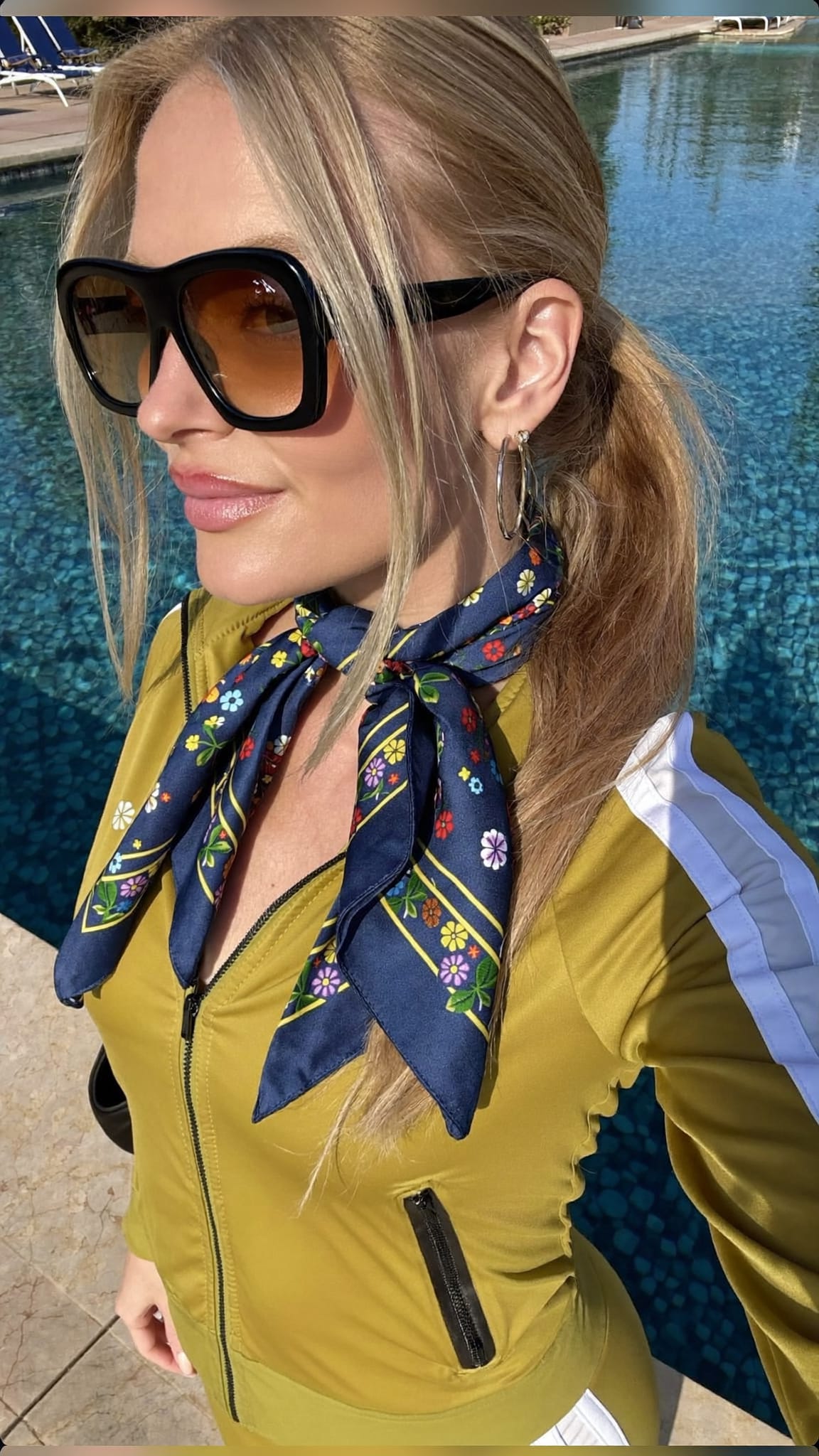 White woman with blonde hair in a low ponytail with a large silk floral scarf tied at the base, wearing oversized sunglasses and a juicy tracksuit at a Beverly Hills pool