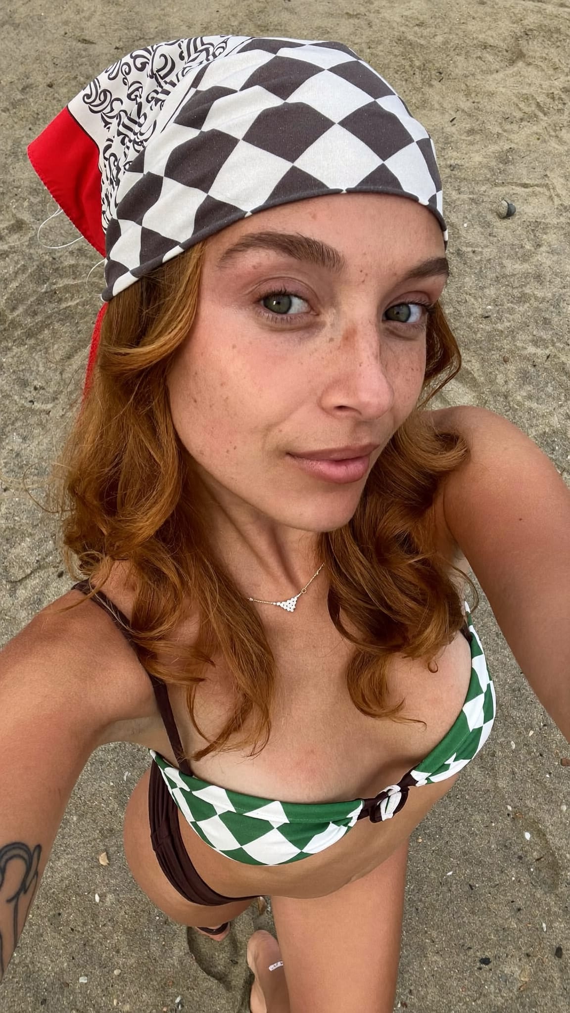 White woman with red hair in tousled beach waves and a printed square bandana worn as a headband, wearing a bikini top and low-rise cutoffs at the beach